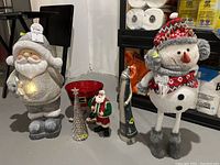 Six holiday decorative figurines including light-up Santa, red bucket, small Santa, light-up tree, large snowman with earmuffs, and snowman on skis.