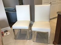 Photo showing two white faux leather dining chairs with silver metal legs side by side on tile floor indoors.