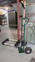 Green metal hand truck dolly with tubular handle and large black wheels next to flat wooden dolly on the floor in basement setting.