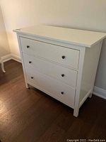 Front view of white wooden dresser with three drawers and black knobs, showing smooth finish and solid build.