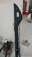 Full view of fishing rod with reel attached leaning against a wall with green fishing rod case behind it