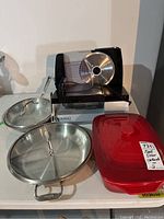 Photo showing Paderno meat slicer with blade and safety guard, two stainless steel covered pans, and a red Pampered Chef casserole dish with lid.