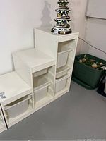 Full view of white storage cabinet showing multiple cubby sections with grey plastic storage bins inserted. Cabinet is against a wall on a basement floor with other items nearby.