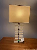 Full view of one table lamp turned on showing the beige fabric rectangular shade and clear acrylic block base.