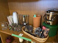 Photo of shelf showing barware lot including vintage drinking glasses, plastic cups and straws, terra cotta wine cooler with label, basket with various drink tools and accessories, and stainless steel ice bucket with green base.