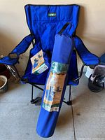 Photo of one blue folding camping chair with armrests and cup holder, accompanied by a matching blue carry bag with product label.