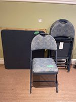 Full view of the folded card table and four folding chairs stacked against a green wall on carpeted floor.