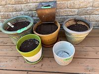 Photo showing six planter pots arranged on wooden surface against brick wall. The pots are varied in size, shape, and color, including ceramic and metal material.