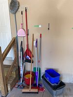 Cleaning supplies including brooms, mops, buckets, bins, and a dustpan grouped by a stairwell.
