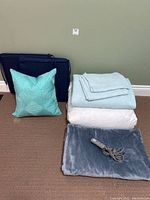Overview of bedding, pillow, cushions, and electric blanket stacked and laid out on floor against wall