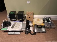 Full view of all items in lot arranged on floor including Sony CD/DVD player, two black Energy speakers, Panasonic landline phone, Sony clock radio, Logitech remote, additional phones, and accessories.