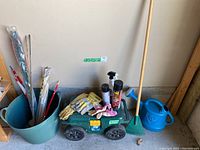 Photo of green garden wagon loaded with gloves, spray bottles, gardening tools including a rake and plant stakes, and nearby blue watering can and bucket