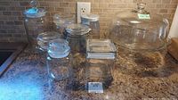 Overview of various glass canisters of different sizes and shapes displayed on a countertop including cylindrical and square shaped containers, some with glass lids and others with metal lids.