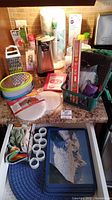 Kitchen items arranged on countertop and drawer including grater, electric can opener, paper towel holder, placemats, napkin rings, tea towels, baking supplies, and gripper pads.