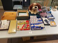Photo showing board games including Rummoli, Rummikub, dominoes, and two stuffed animals placed on a table.