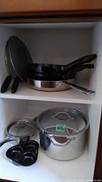 Image of various pots, pans, and splatter screens in white cabinet shelves. Showing handles and lids.