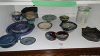 Photo showing various pottery items including bowls, dishes, a utensil holder, and decorative jar with floral motif, arranged on a white surface.