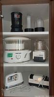 Shelf view of the kitchen appliances showing Magic Bullet, Black+Decker steamer, Cuisinart Mini-Prep chopper, hand mixer, iron, and Chef's Choice knife sharpener
