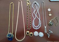Top view of all jewelry items laid out including necklaces, earrings, charm, and pocket watch with chain on a wood surface