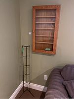 Wall mount wood display cabinet with glass door closed, hanging on wall above carpeted floor near couch, with metal rack standing beside it.