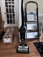 Photo showing a Bissell Crosswave cleaner and its plug-in charging stand. There is a cardboard box on the floor nearby and a black and white floor mat partially visible.