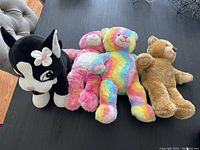 Four plush toys on a couch: one black and white puppy, a pink bear, a rainbow-colored bear, and a brown bear.