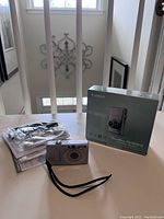 Canon PowerShot SD1200 IS camera on white surface with original box and accessories including cables and manuals shown
