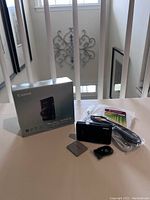 Lot contents shown from front including Canon box, camera, remote control, camera stand, charging cable, manuals, wrist strap, and sealed package of a Transcend 16GB SD card (not included).