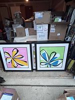 Two custom double-framed colorful flower artworks resting on the floor among boxed items. Each art piece shows large abstract flowers with bright petals and strong black outlines.