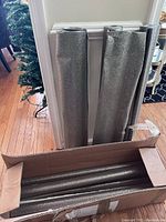 Two rolls of glitter wallpaper resting against a wall near a decorative Christmas tree, inside and out of the cardboard box they came in, showing their reflective textured surface.