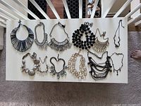 Top view of 12 fashion necklaces arranged on white surface showing variety in size, design, and materials. Includes black reversible necklace and several with rhinestone sparkle elements.