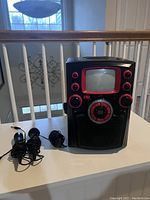 Front and angled view of karaoke machine with black and red accents, showing screen, control knobs, and speaker grille. Two wired microphones and charger positioned beside machine.