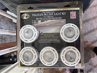 Front view of unopened Halogen Accent Light Kit packaging showing five white circular puck lights arranged in two rows, with mounting instructions and hardware visible.