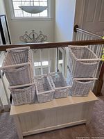 Photo showing 6 white wicker baskets arranged on a white storage bench; 4 taller baskets on the edges, 2 smaller baskets in the middle.