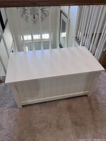 Closed white wooden storage chest with vertical panel design on sides and front, placed on carpet near stair rails.