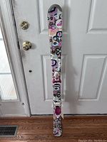 Full-length image of a single ski with white base and colorful flower graphics in pink, purple, brown, black, and blue. The ski has attached bindings. Placed upright against a door for perspective of size and design.