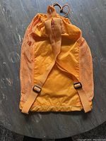 Back view of orange mesh drawstring backpack with adjustable straps laying flat on wood floor.