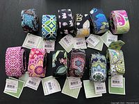 Top-down view of 12 rolled Vera Bradley cloth belts with original tags displaying various colorful patterns.