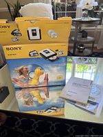 Photo shows the Sony DVDirect device in original box and packaging, along with manuals and DVDs on a glass table with background of a window and indoor plants.