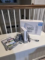 White Nintendo Wii console upright with attached power cord, original box behind, and a small stack of Wii game cases in front.