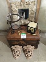 Photo showing decorative wooden box with metal leaf detail, framed mirror in an ornate cream frame, two carved corbels, metal candle holder with glass insert, square glass candle holder with candle inside, and candle snuffer