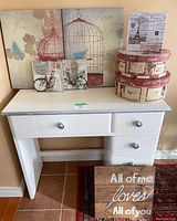 White painted wooden desk with silver trim on edges, showing all drawers and top surface with decor items placed on it, including canvas art and hat boxes.