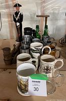 Photo showing assorted 13 beer steins with different designs including coats of arms and wildlife, 2 green Grolsch bottles, a decorative figure statue, and some wooden keg accessories on a wooden shelf.