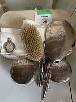Photo of international sterling vanity set including hand mirror, brush, and two containers along with white gloves and pocket watch.
