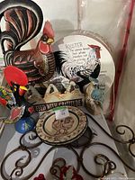 Photo showing various rooster-themed items including two plates, wooden rooster figurine, wooden parrot, rooster hat box, and decorative sign arranged on a surface.