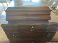 Photo showing stacked view of two wooden jewelry boxes. Larger box on bottom has three brass handles; smaller box on top has a flat lid.