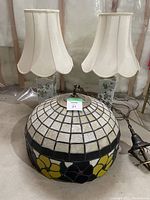 Stained glass dome-shaped hanging lamp with grid pattern on top and band of yellow and purple flowers on the bottom, placed on basement floor with attached hanging fixture and cords