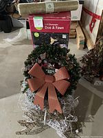 Photo showing Champagne Doe & Fawn box, green wreath with red plaid bow, and some white wire decoration on floor.