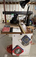 Wide shot of Craftsman 10-inch radial arm saw mounted on a thick wooden table with visible ruler marks, with extra blades hanging on the side pegboard, extension cords coiled below, and large red dustpan attached below.