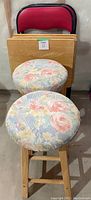 Photo of two wooden stools with round floral cushioned seats and a folding chair with red fabric and black trim behind.
